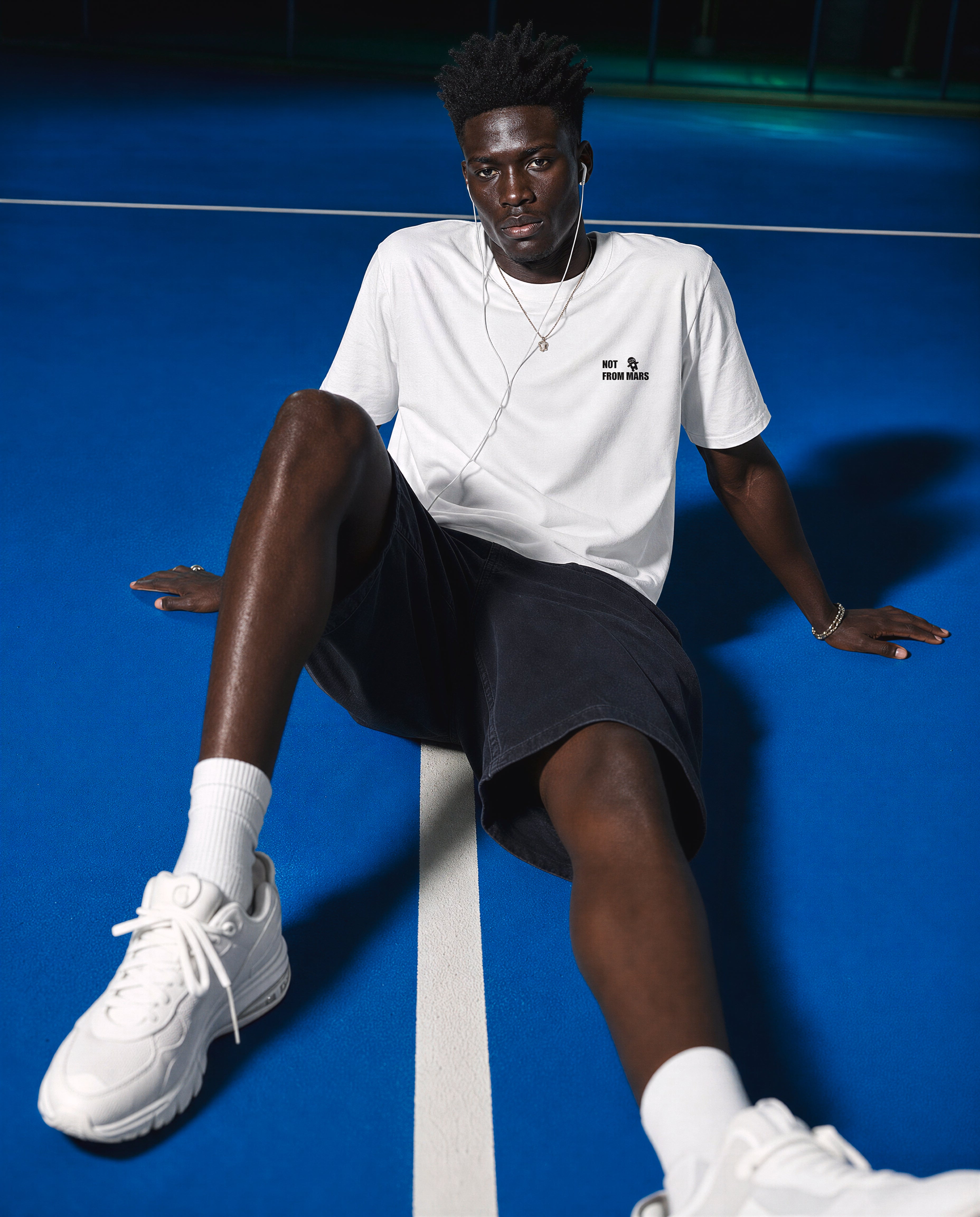 A man sits on a blue tennis court wearing a white short-sleeve t-shirt with a small black logo on the left chest, black athletic shorts, white socks, and white sneakers.